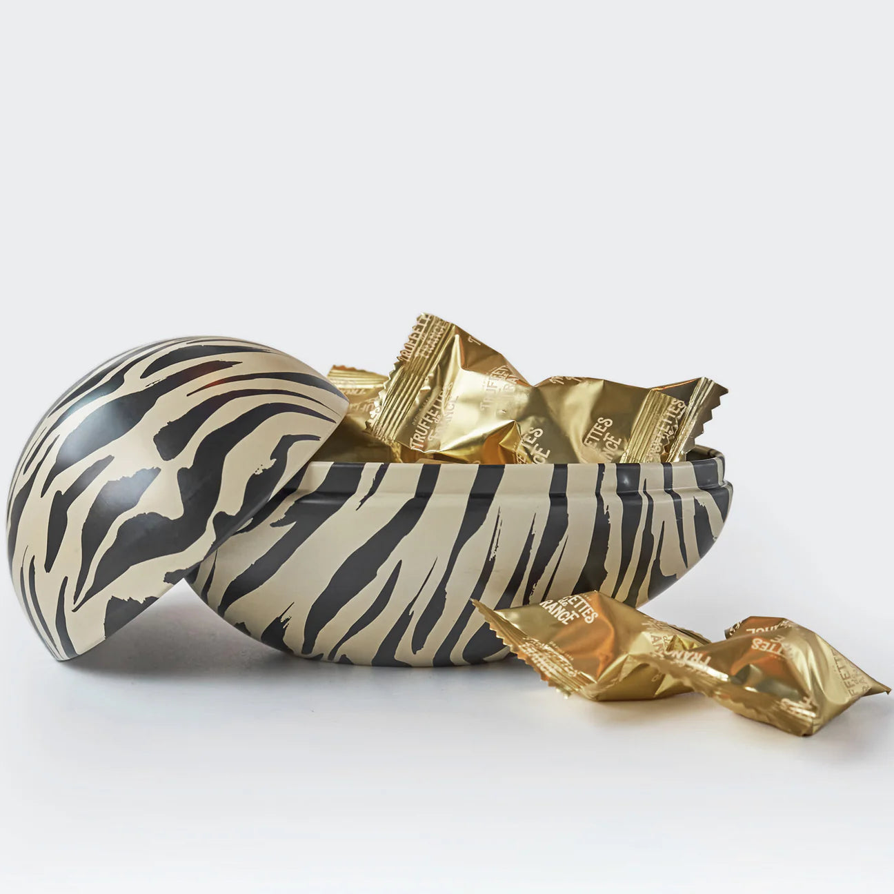 The Gåva Gifting Tiger Reusable Easter Egg, a Swedish-style Påskägg in tin with black and beige zebra print, holds several gold-wrapped candies. Its lid rests to the side, with two treats placed outside on a white surface.