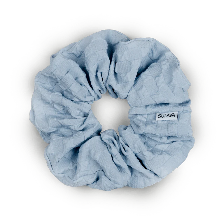The Daydream Scrunchie in Blue by Sui Ava features a textured light blue design and a white label with the brands name, offering a gentle elastic that brings a dreamy touch to your hairstyle.