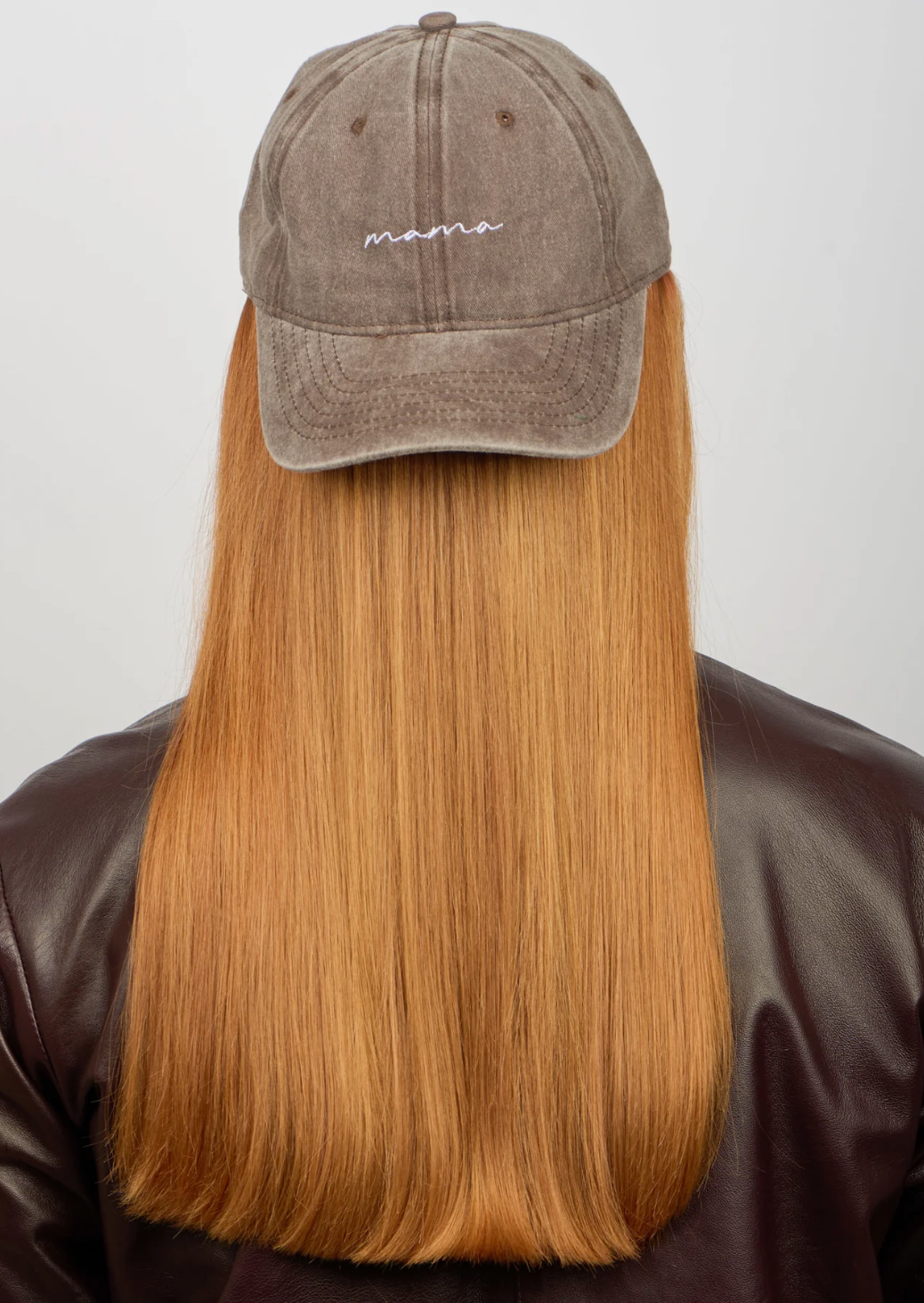 A person with straight, long, reddish-blonde hair wears the Sui Ava Everyday Cap, Mama—a washed brown adjustable cotton cap with "mama" embroidery—and a dark brown leather jacket, photographed from behind against a plain background.