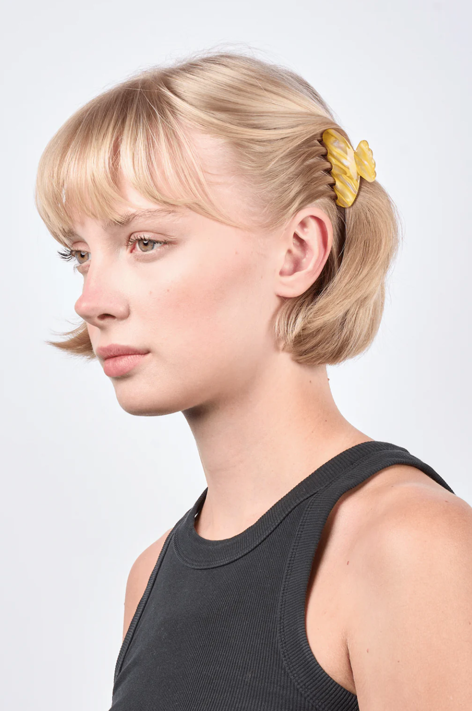 A woman with short blonde fine hair wears a black sleeveless top and accessorizes the left side of her head with the Helle Fresh Mini Hair Claw Clip in Yellow by Sui Ava, shaped like pasta.