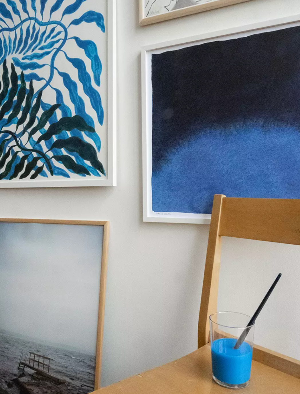 Colorful wall art featuring deep blue abstract posters and a glass of blue paint on a wooden chair.