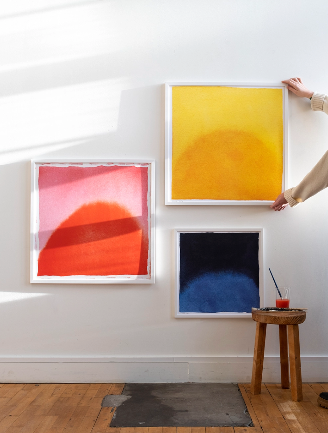 A hand adjusts the Sun Print (70x70 cm) by Fine Little Day, one of three framed abstract artworks on a white wall. A stool with a drink and paintbrush nearby completes the creative scene.