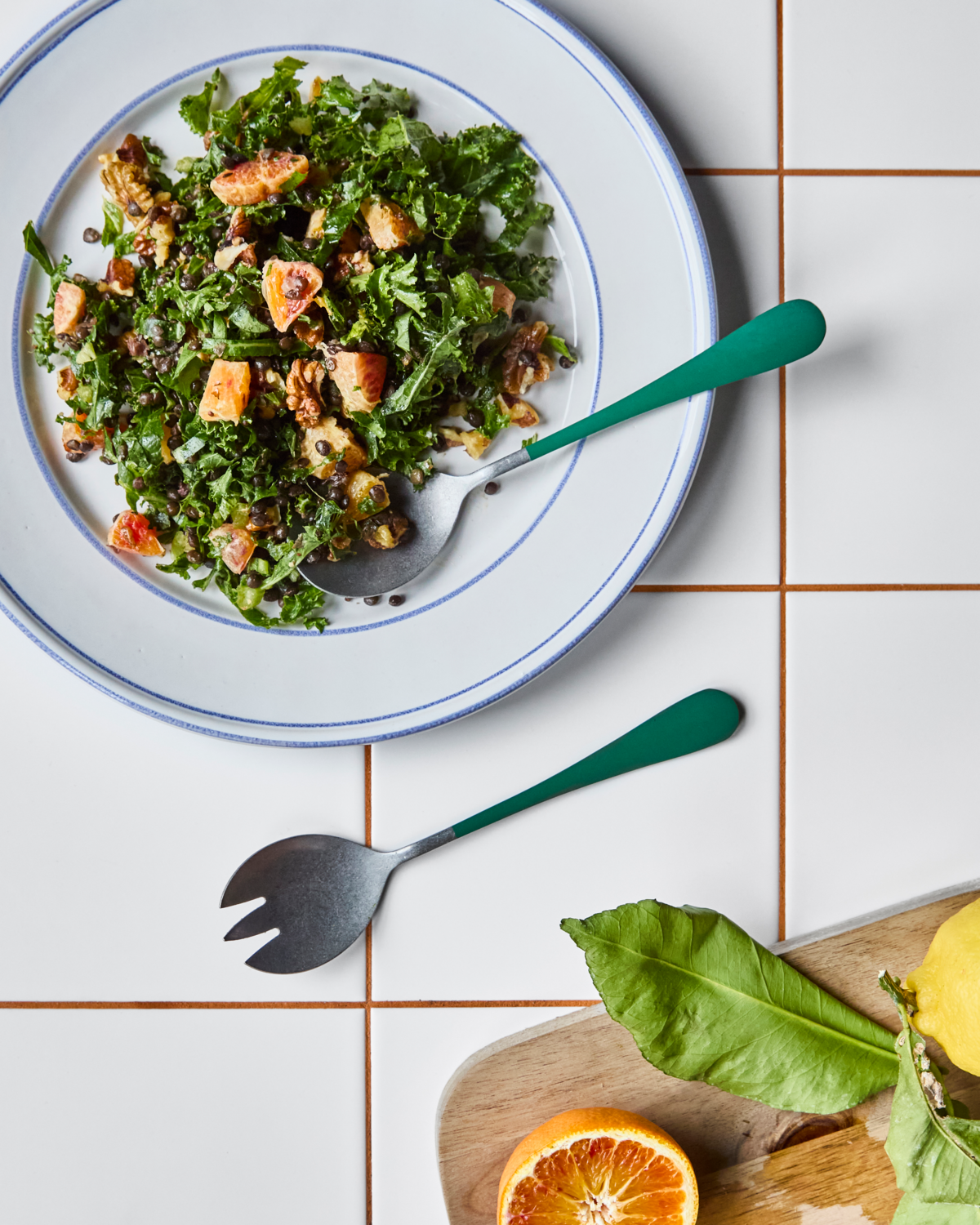 A plate of kale salad with fruits and nuts is served on a white tiled surface, paired with the Nicolas Vahé Salad Set in Stainless Steel, Green—ideal for elegant dining. A cutting board nearby holds a lemon, leafy greens, and half an orange.