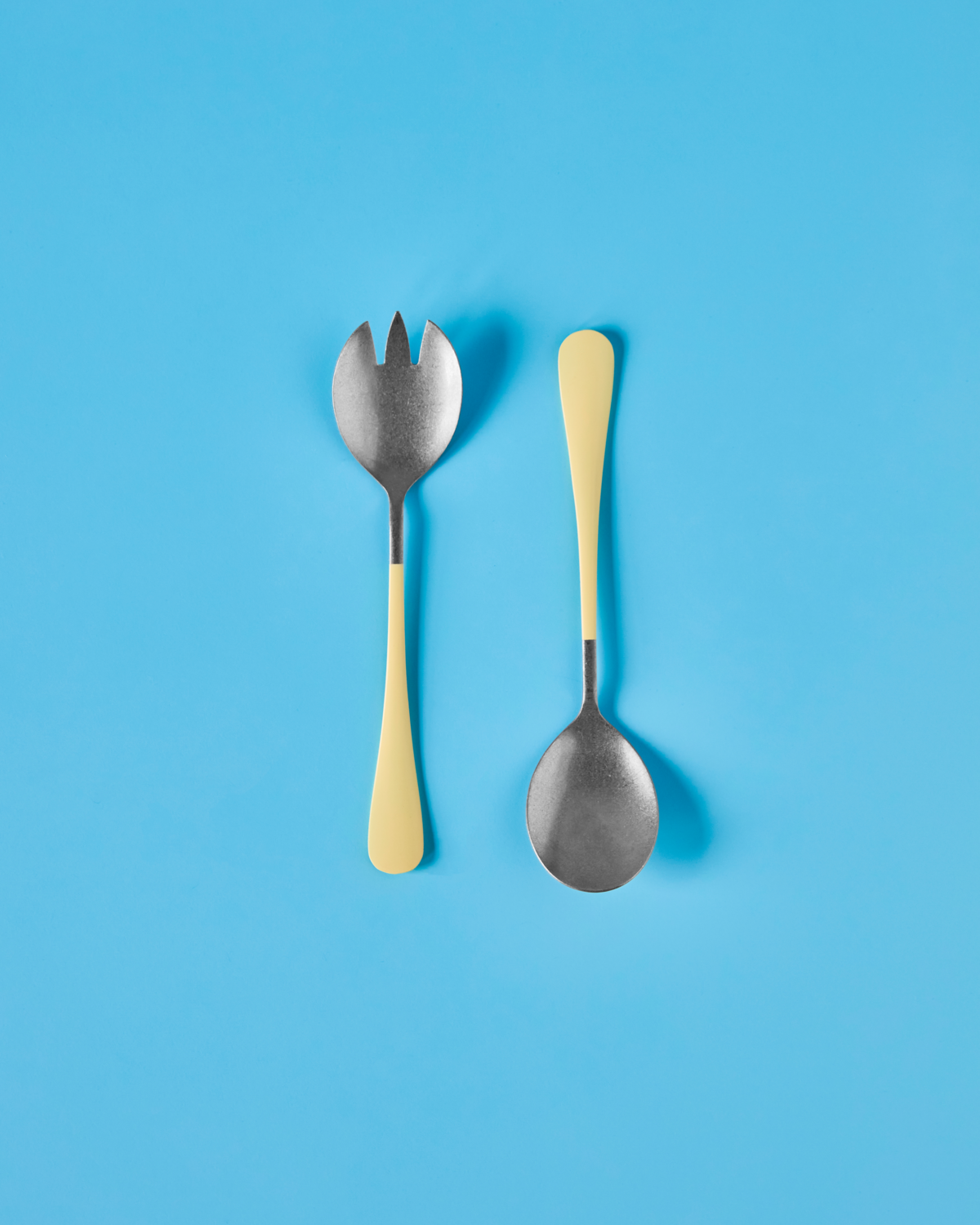 The Nicolas Vahé Salad Set in Stainless Steel with yellow handles is displayed side by side on a bright blue background.