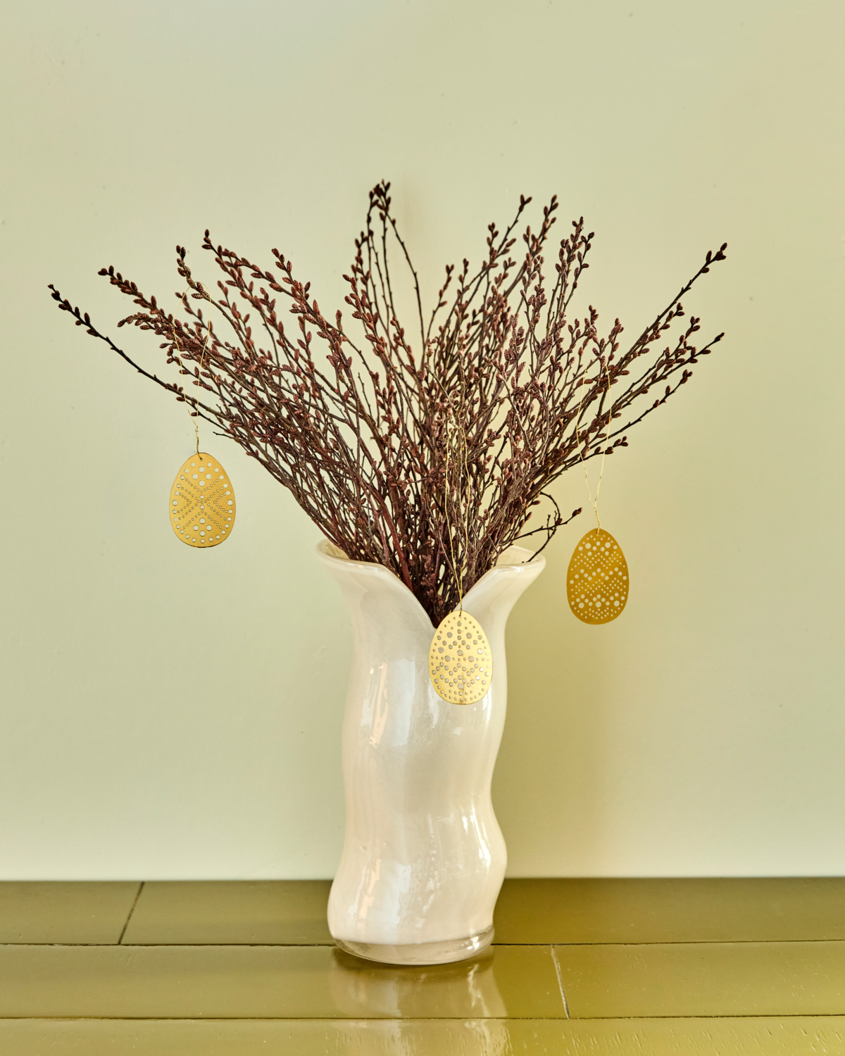 A white ceramic vase holds dried brown branches decorated with House Doctor's Easter ornaments— a set of 3 cut brass eggs—arranged on a green surface against a pale green wall.