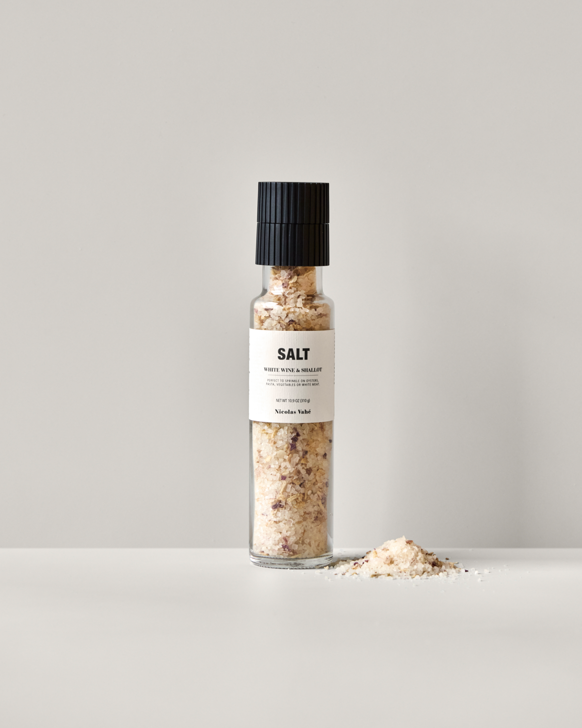 A clear glass grinder labeled "Nicolas Vahé Salt, White Wine & Shallot" sits on a light surface with some coarse sea salt blend spilled in a small pile beside it.