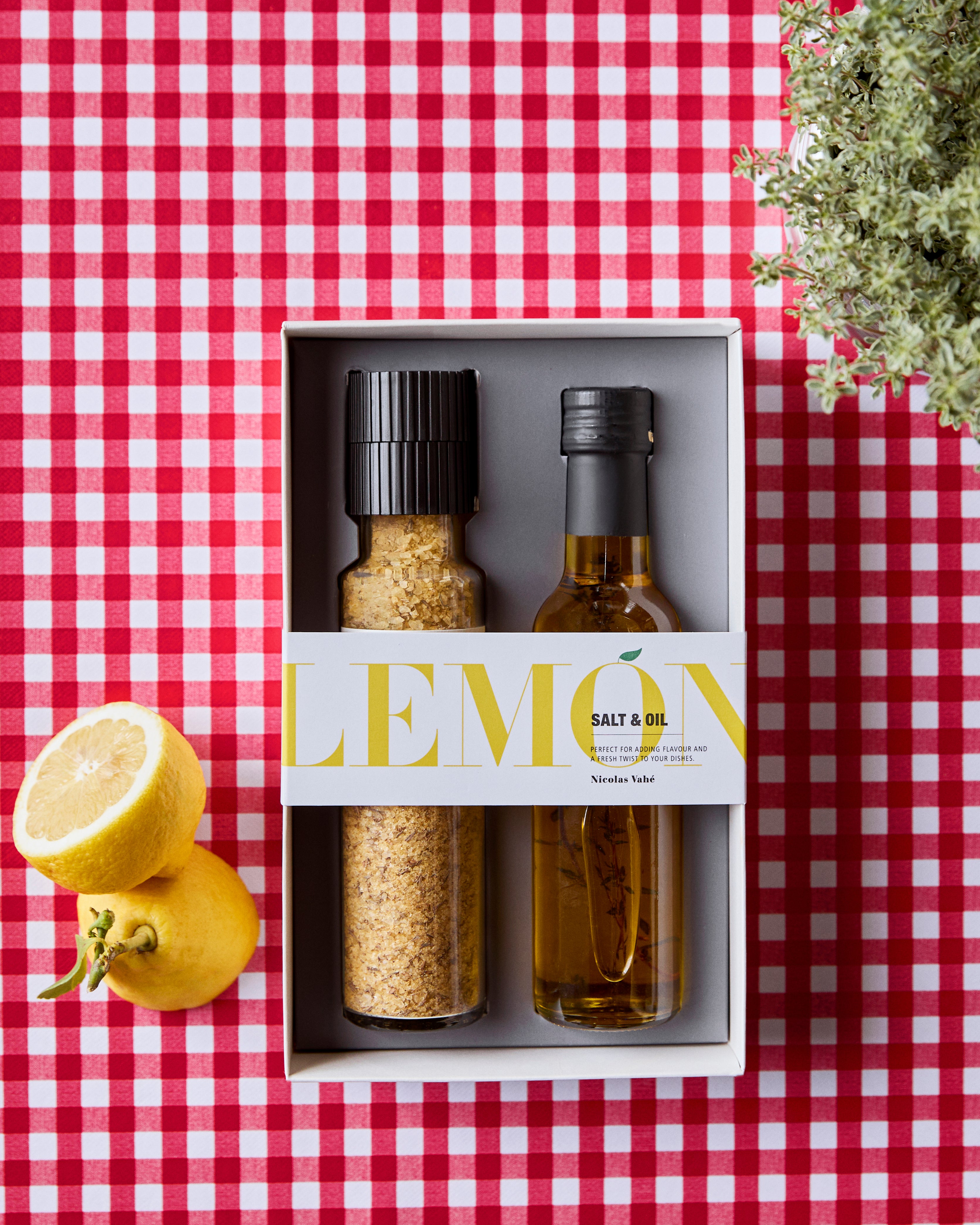 A red checkered tablecloth holds the Salt & Oil Lemon, Gift Box from Nicolas Vahé, which includes a lemon and thyme seasoned salt grinder and a bottle of lemon and thyme oil. The box is labeled "LEMON" and is placed beside two fresh lemons and a leafy plant.