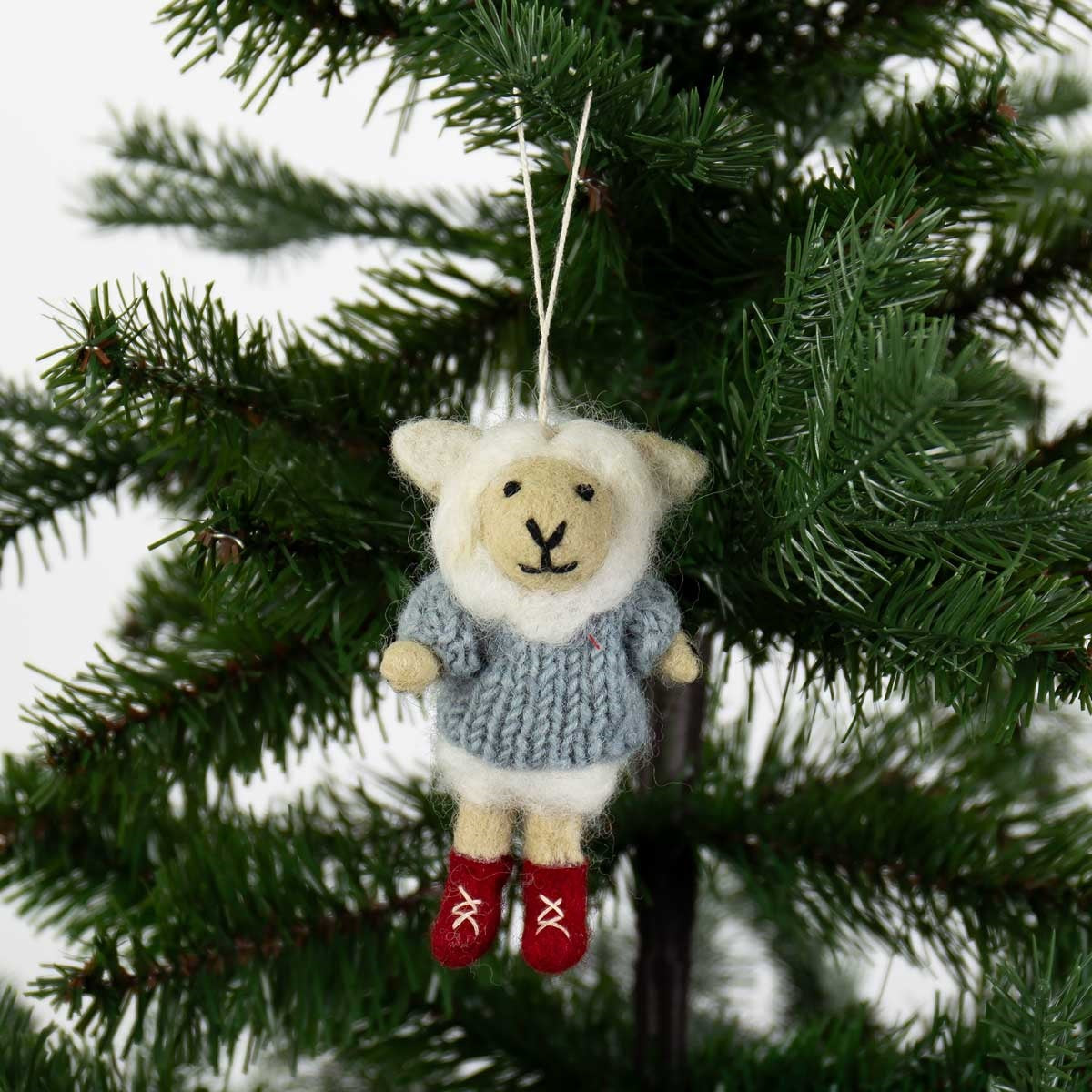 A Sheep in Red Boots Christmas Ornament, Handmade in Wool, featuring a felt sheep in a gray knitted sweater and red boots, hanging from a holiday tree branch.