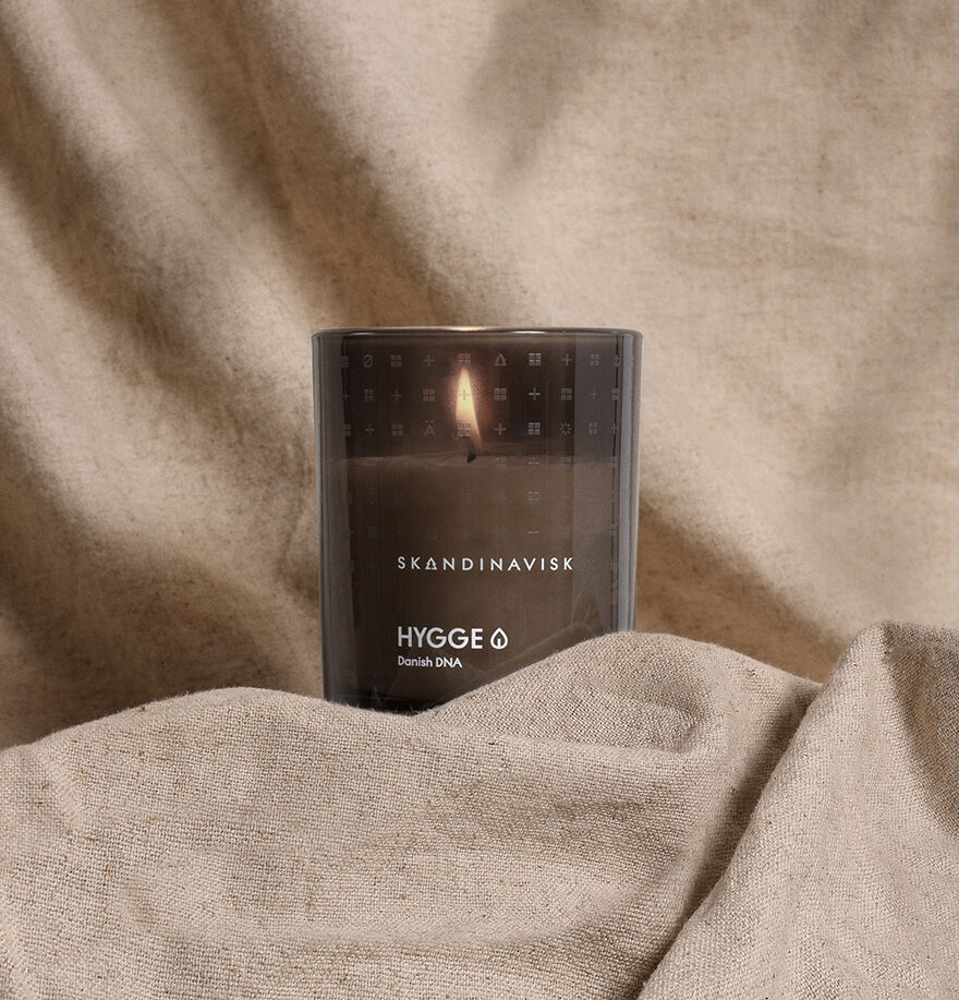 The Skandinavisk Scented Candle Hygge 50h, 200g glows warmly on beige fabric with a neutral background, embodying the cozy essence of Scandinavian comfort.