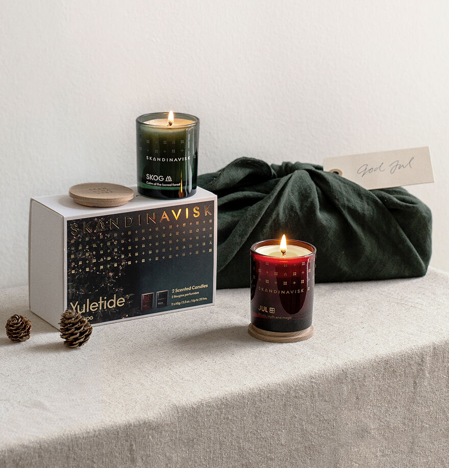 The Skandinavisk Giftset Yuletide features Jul and Skog mini scented candles—one lit, one unlit—styled with a gift box, dark green cloth wrap, labeled tag, and pine cones on neutral fabric for an ideal seasonal candle display.