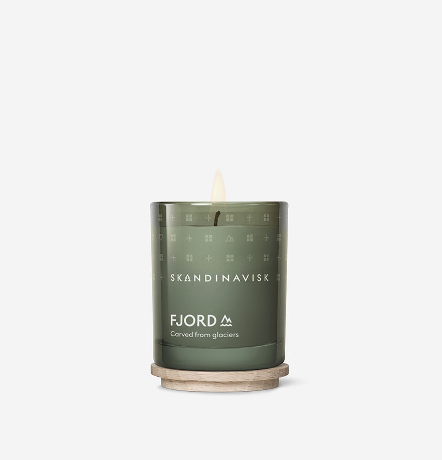 A lit Mini Scented Candle Fjord 20h, 65g by Skandinavisk, crafted from Swedish rapeseed wax, sits on a round wooden coaster with its engraved beechwood lid nearby, all set against a plain white background.