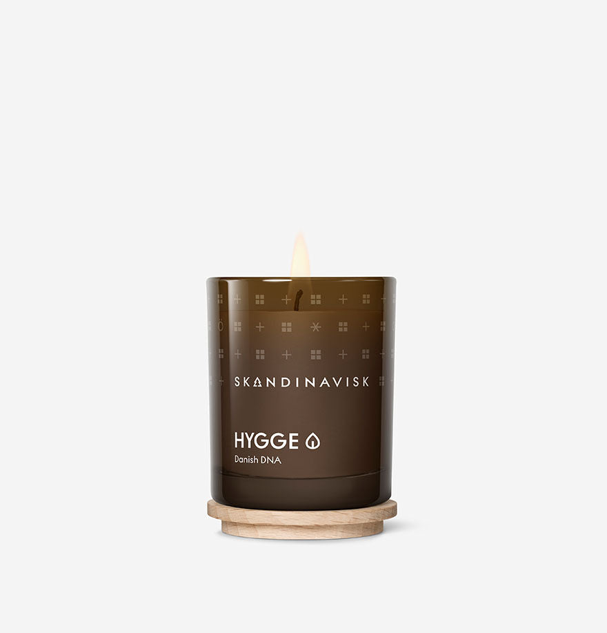 The Skandinavisk Mini Scented Candle Hygge 20h, 65g glows in a dark glass holder with a wooden base, made from Swedish rapeseed wax, and is photographed against a white background.