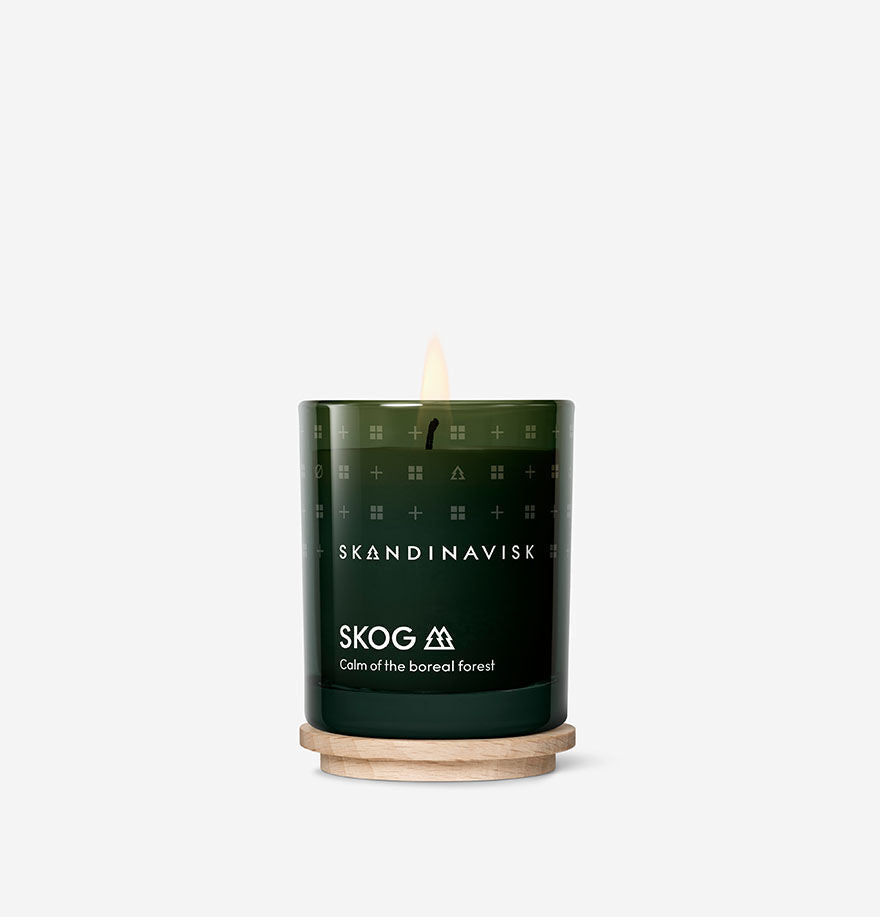 A lit Mini Scented Candle Skog 20h, 65g by Skandinavisk, inspired by the boreal forest, rests on a wooden base against a plain white background.