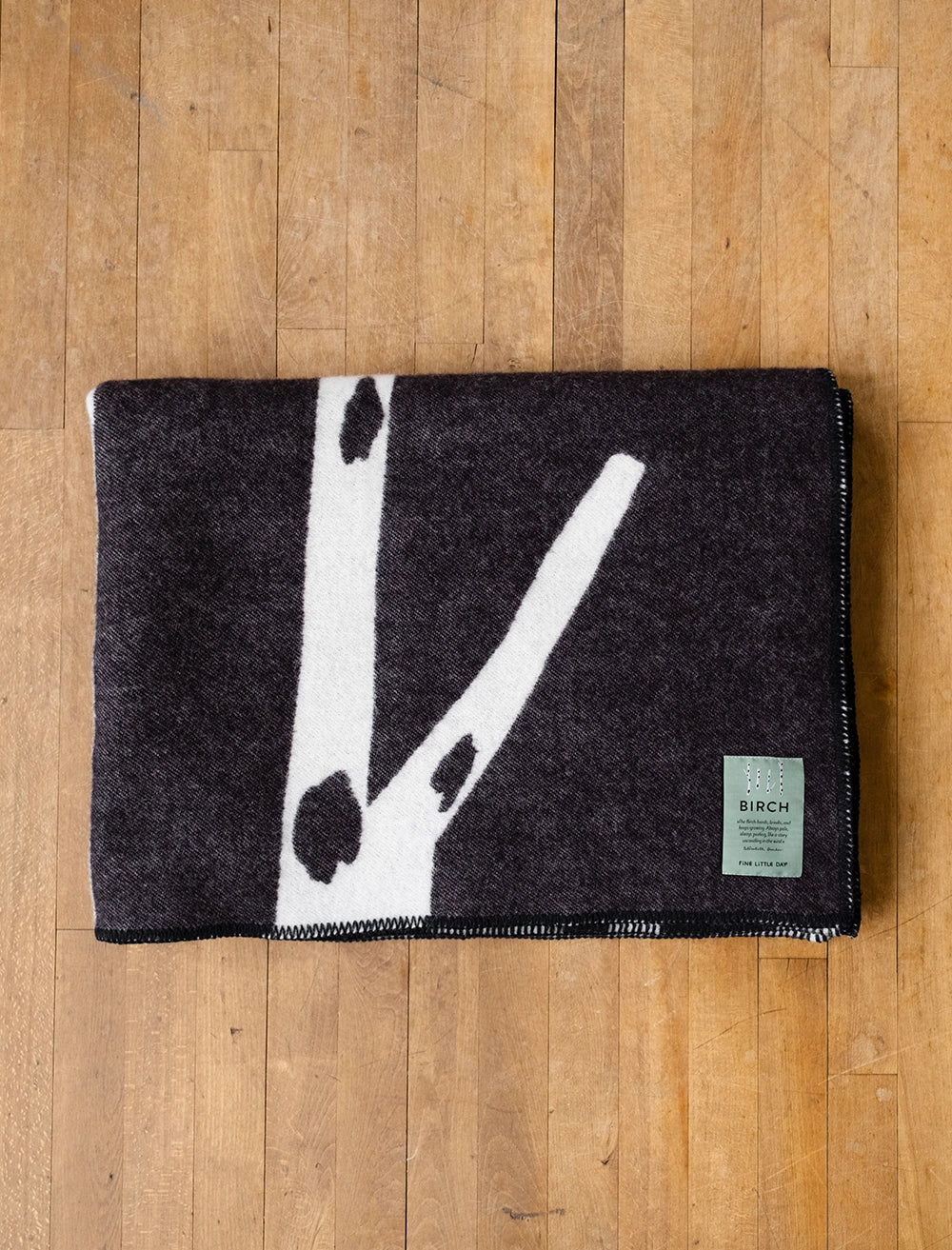 The Fine Little Day Birch Wool Blanket, reversible in black & white, is folded on a wooden floor with a green label at the bottom right corner.