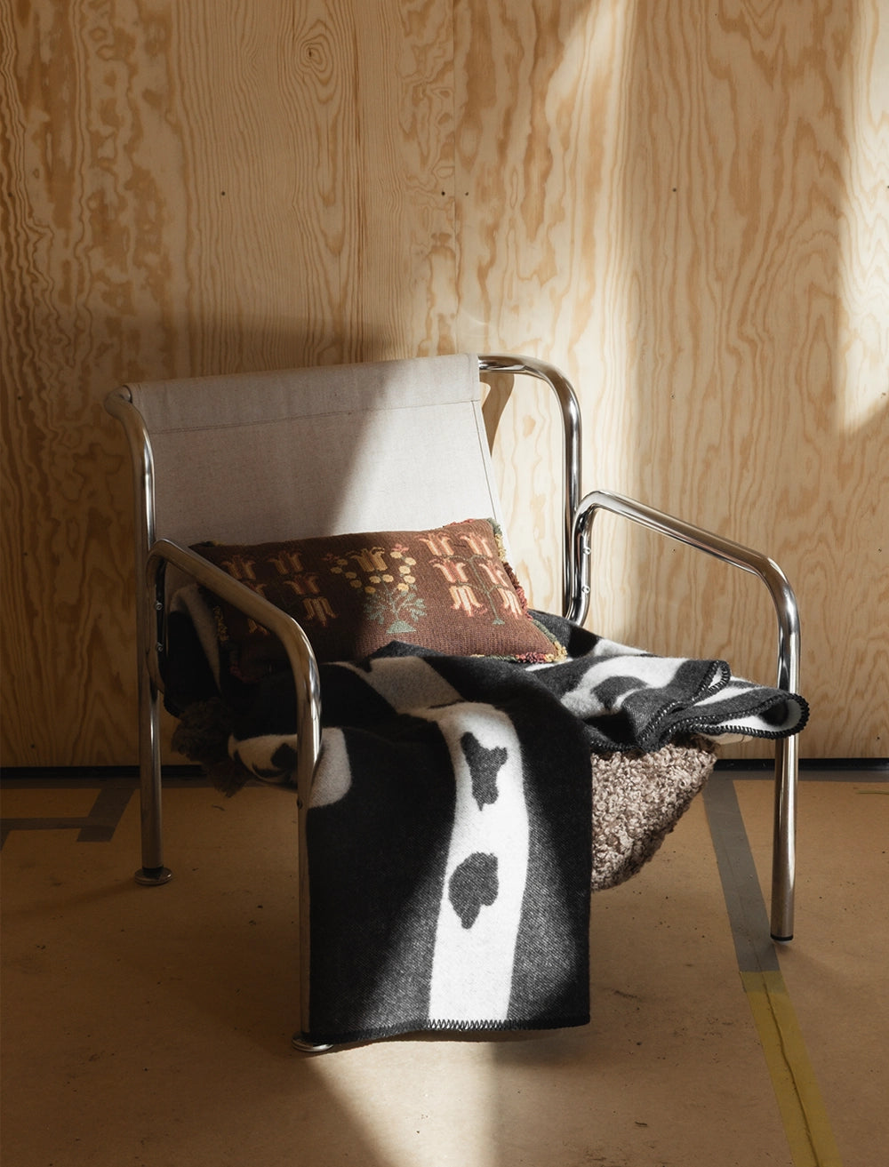 A metal-framed chair with a light fabric seat features a patterned cushion and the Fine Little Day Birch Wool Blanket, Reversible in Black & White, all arranged against a light wood-paneled wall inspired by Scandinavian design.