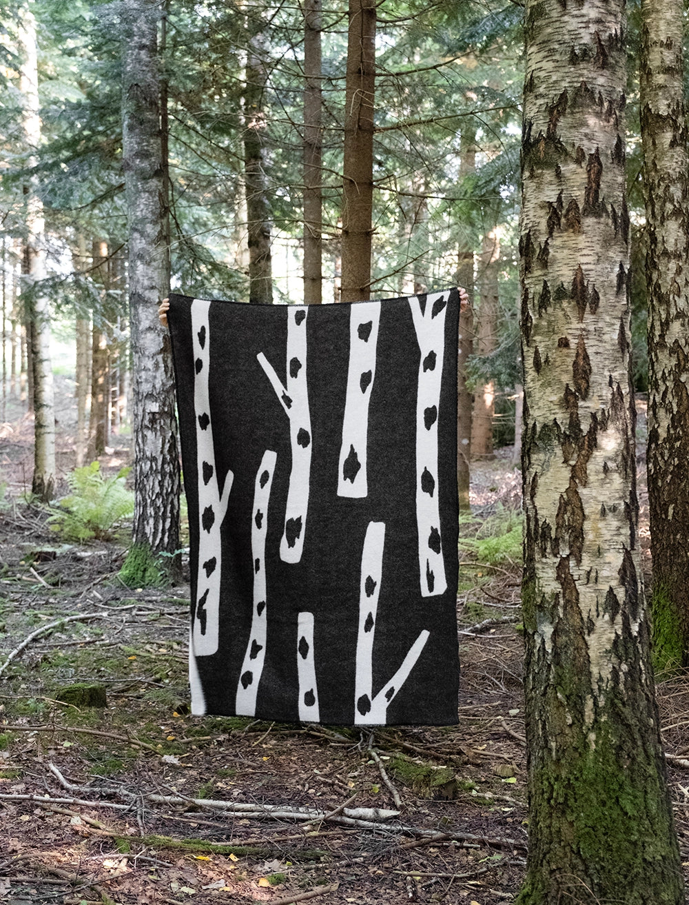 The Birch Wool Blanket, Reversible in Black & White by Fine Little Day hangs vertically among forest trees, adding Scandinavian style to the natural setting with its distinctive birch pattern.