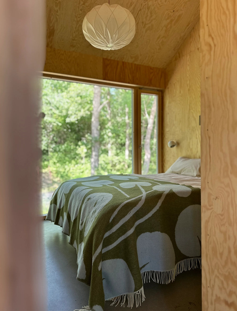 A simple bedroom with wooden walls, a large window framing green trees, and a bed topped with Fine Little Day's Water Lilies Wool Extra Large Blanket/Bedspread in reversible blue and green.