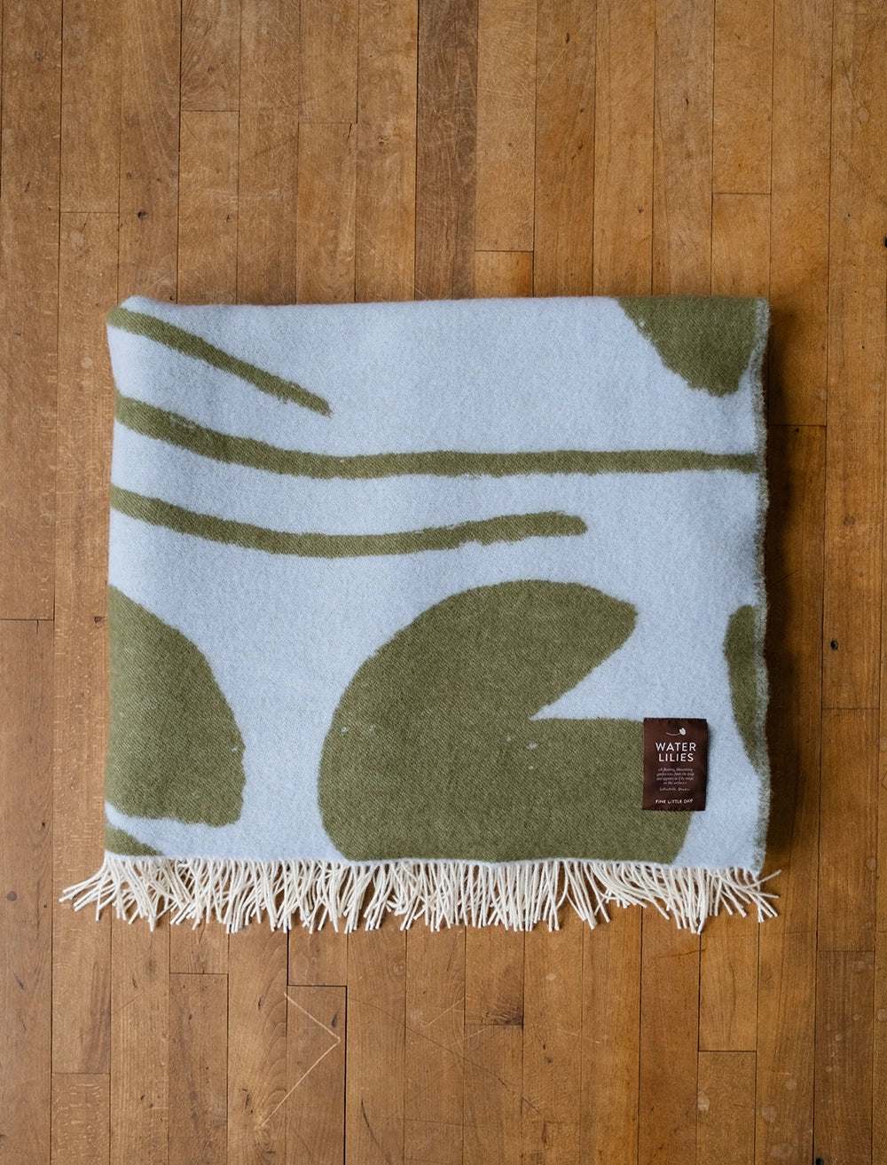 A Fine Little Day Water Lilies Wool Extra Large Blanket / Bedspread in blue & green, reversible with fringed edges, rests folded on a wooden floor. The brand label is visible on one corner of the woven blanket.