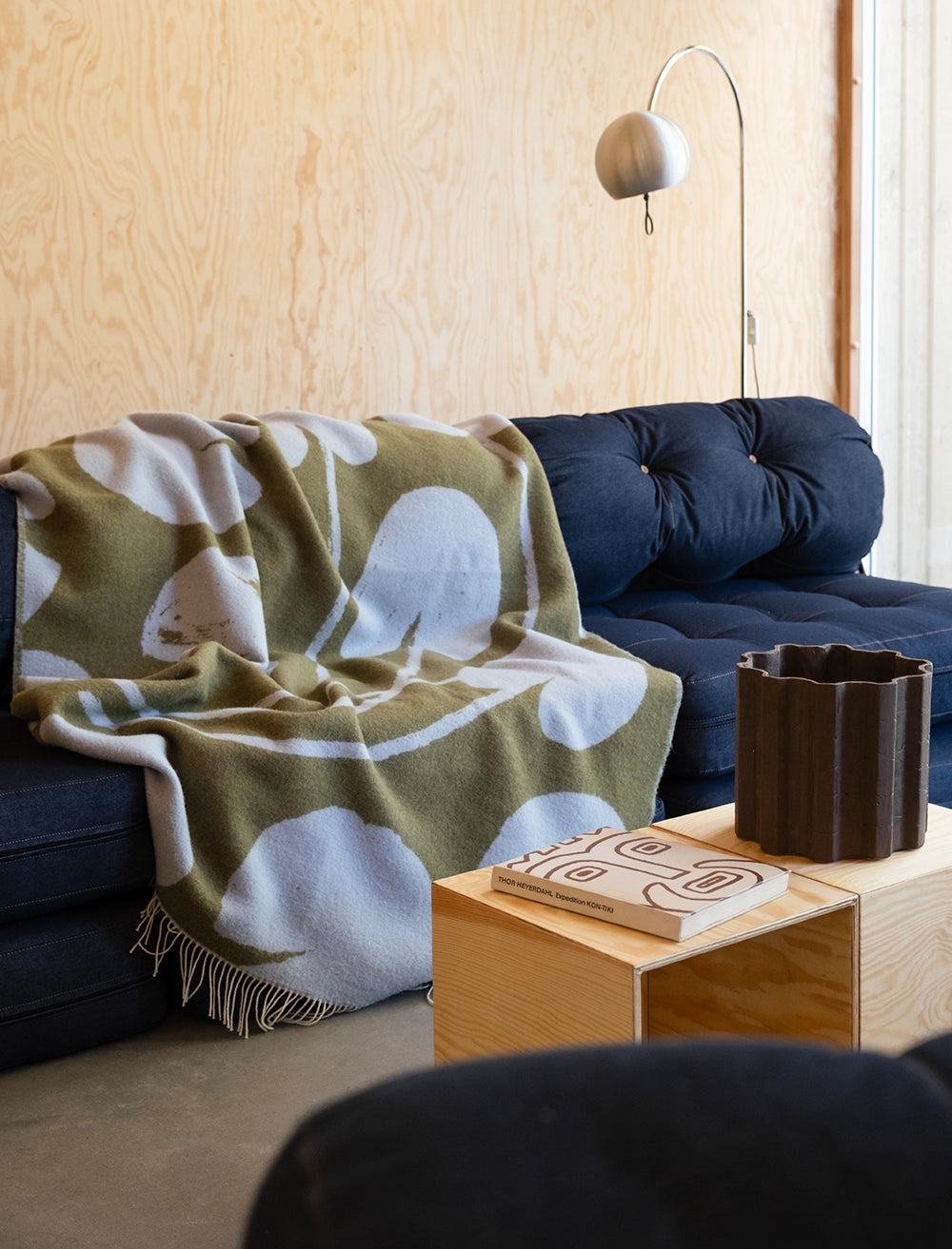 A dark blue sofa with the Fine Little Day Water Lilies Wool Extra Large Blanket, reversible in blue and green, a wooden coffee table holding two books, and a dark vase in a modern wood-paneled room.