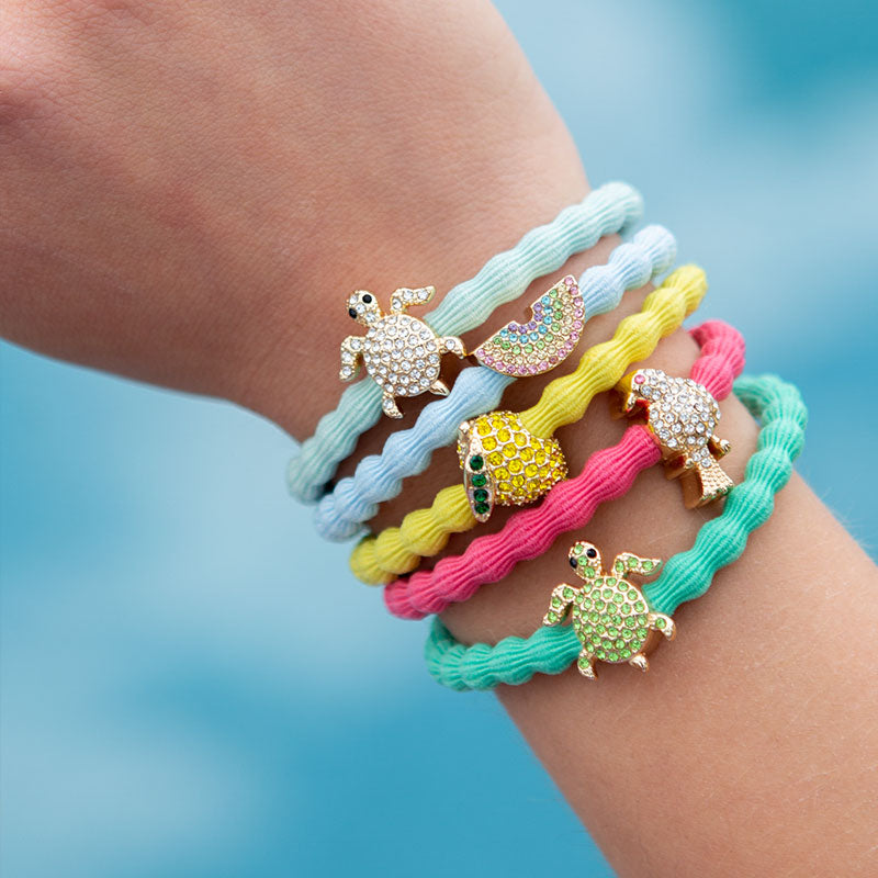 A hand displays five ByEloise pastel-colored bracelets, including the Mint Bangle Band Turtle, each featuring a jeweled animal charm—turtle, rainbow, parrot, pineapple, and frog—adorned with sparkling diamanté. A blurred blue background evokes the look of water.