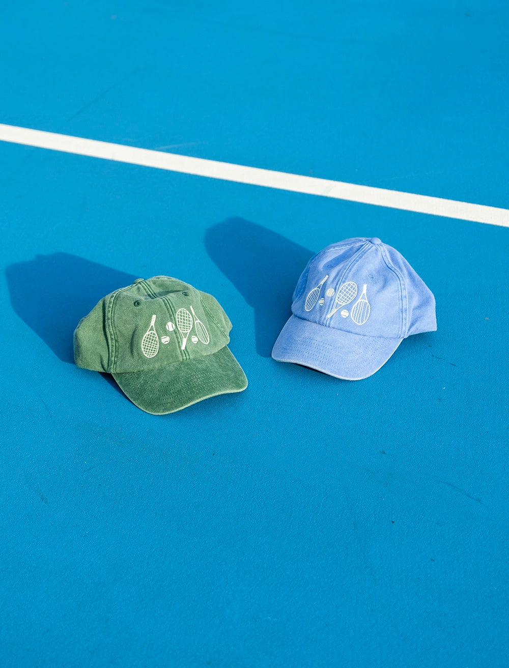Two Fine Little Day Tennis Caps in Vintage Green and Light Blue, both embroidered with tennis rackets and featuring adjustable straps for a perfect fit, lie on a bright blue court by a white line.
