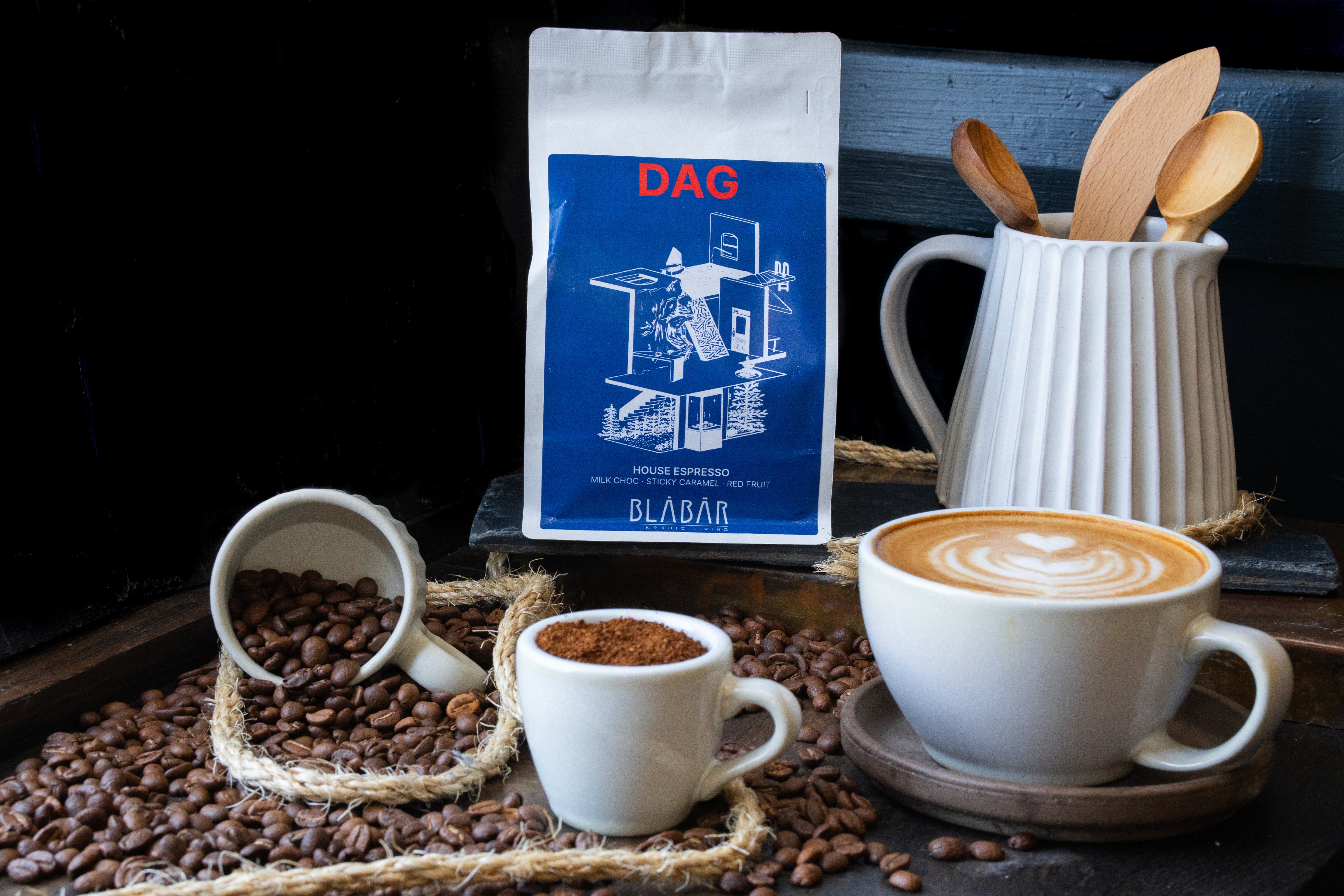 A cup of cappuccino with latte art, a white cup of rich espresso coffee, a spilled cup of coffee beans, and a bag of Blåbär Nordic Living DAG House Coffee Beans Blåbär, Single Origin Colombia, 250g are arranged on a wooden surface next to a jar holding wooden utensils.