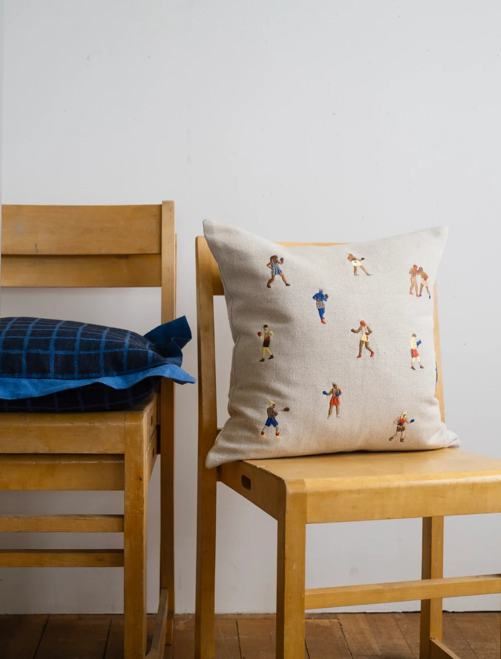 Two wooden chairs sit side by side; one has a blue checkered cushion, and the other features Fine Little Day’s Boxers Embroidered Cushion Cover, decorated with small, colorful figures. A plain white wall forms the background.