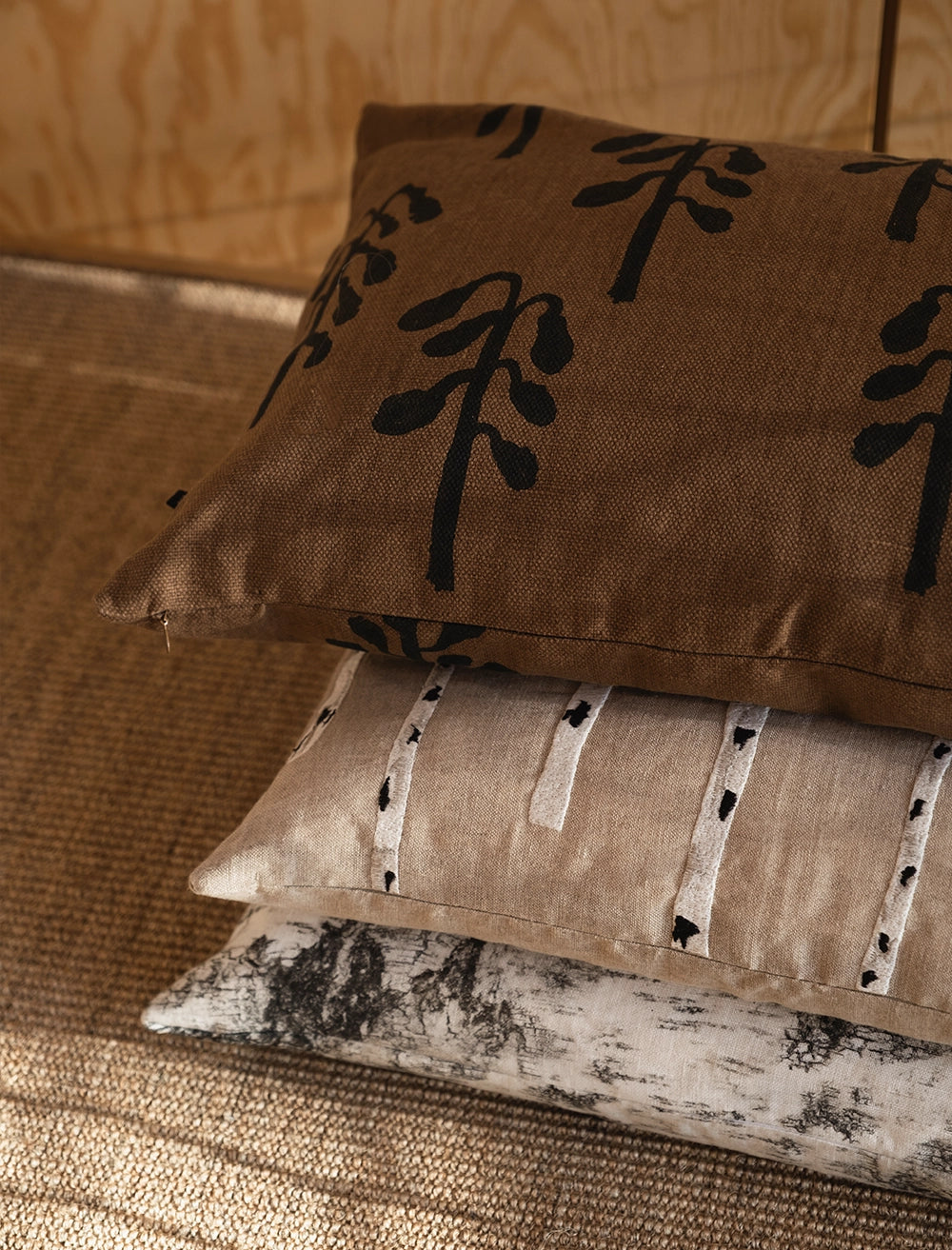 Three decorative pillows, including the Birch Embroidered Cushion Cover in Nature by Fine Little Day, are stacked on a textured brown surface with a wooden background.