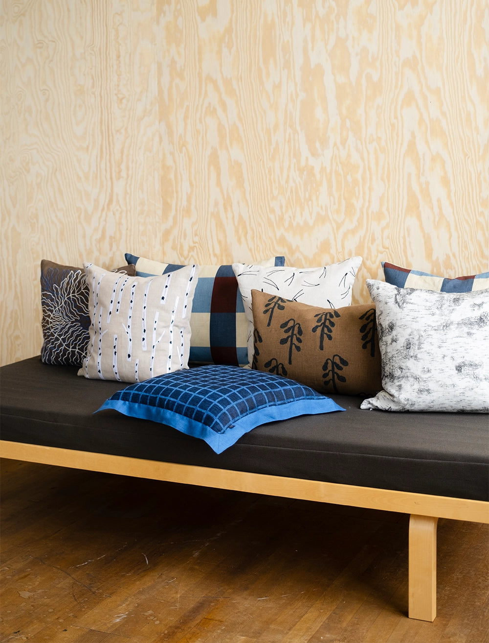 A wooden daybed with a dark cushion is topped with seven pillows, including the Birch Embroidered Cushion Cover in Nature by Fine Little Day, featuring blue, brown, white, and black patterns against a light wood wall.