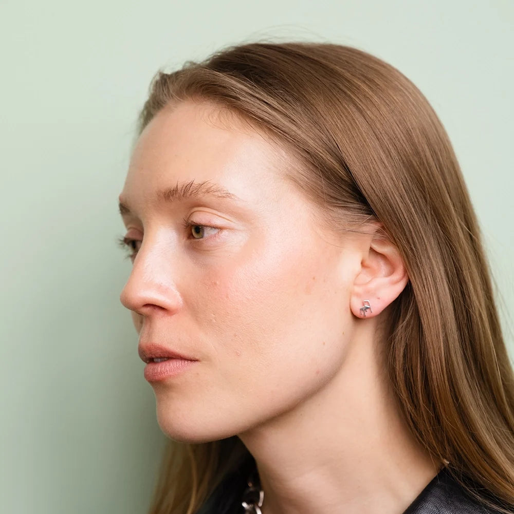 A woman with long, light brown hair and clear skin is shown in profile against a pale green background, wearing the Chair Stud Earrings in Silver by Fine Little Day—small recycled silver earrings inspired by the elegant lines of Orchestra Chair jewelry—and a dark top.