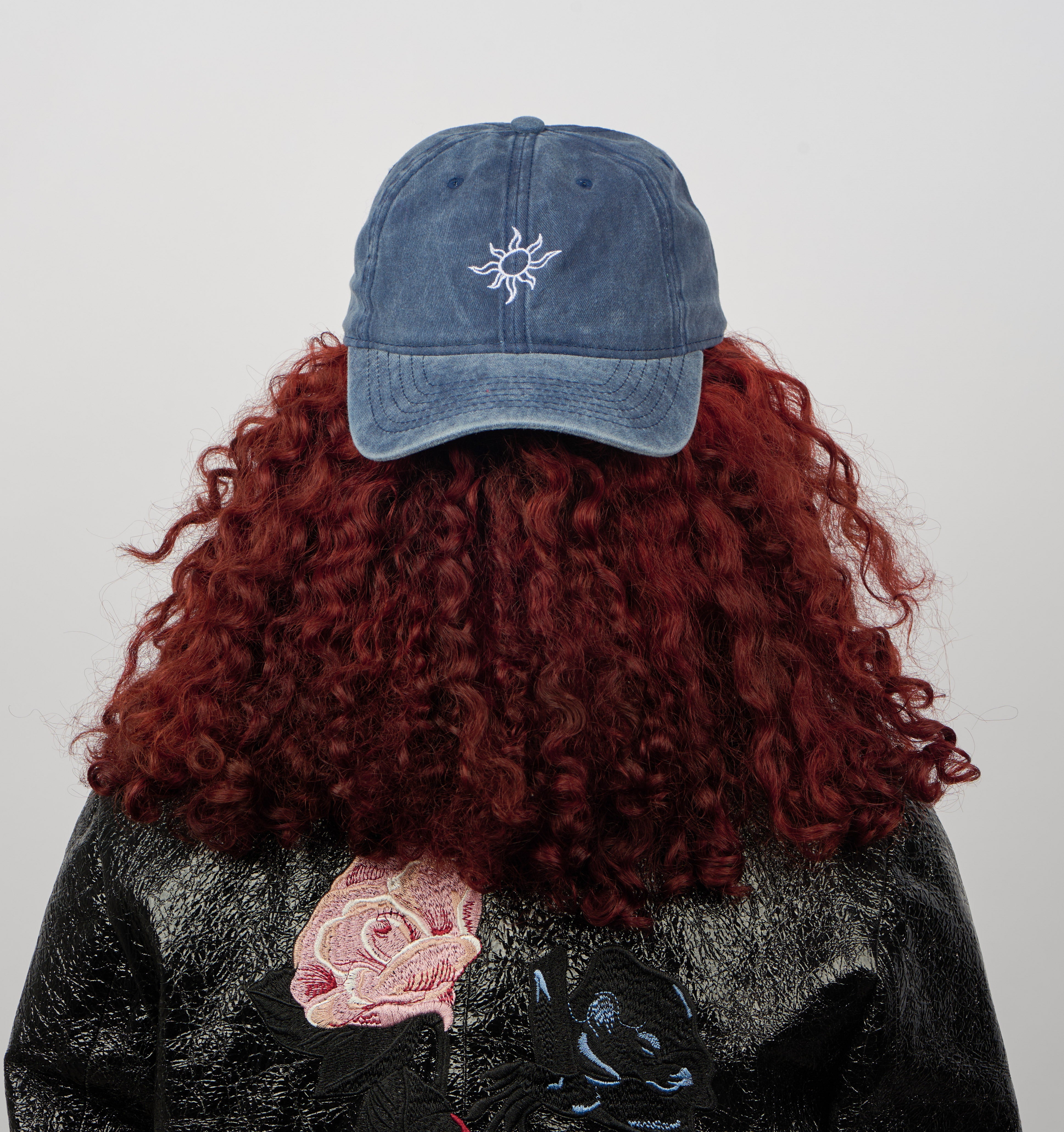 A curly red-haired person, seen from behind, wears the Sui Ava Everyday Cap in White Sun/Faded Blue with an adjustable strap and a black jacket embroidered with flowers and animals. The plain background is light-colored.