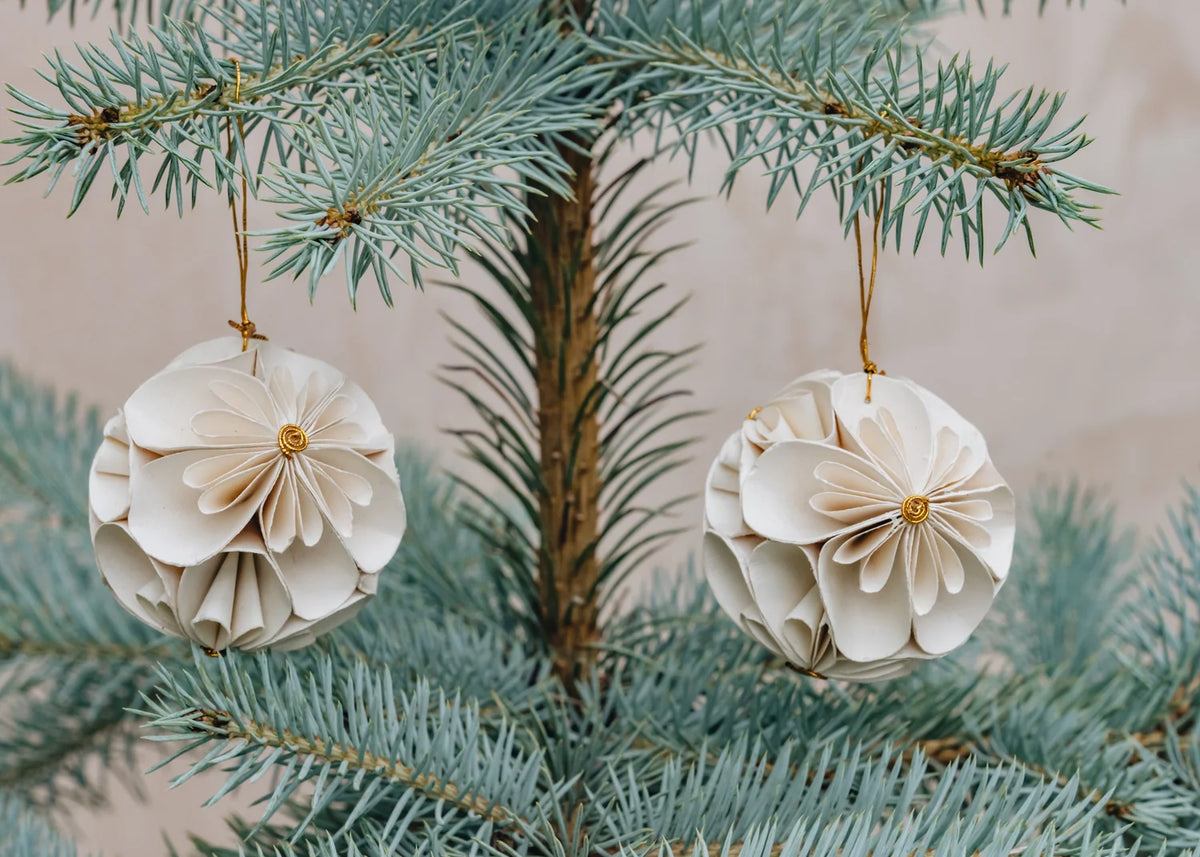Two handmade Flower Ball Christmas Ornament 2-pack in White spherical paper flowers hanging from the branches of a blue spruce tree.