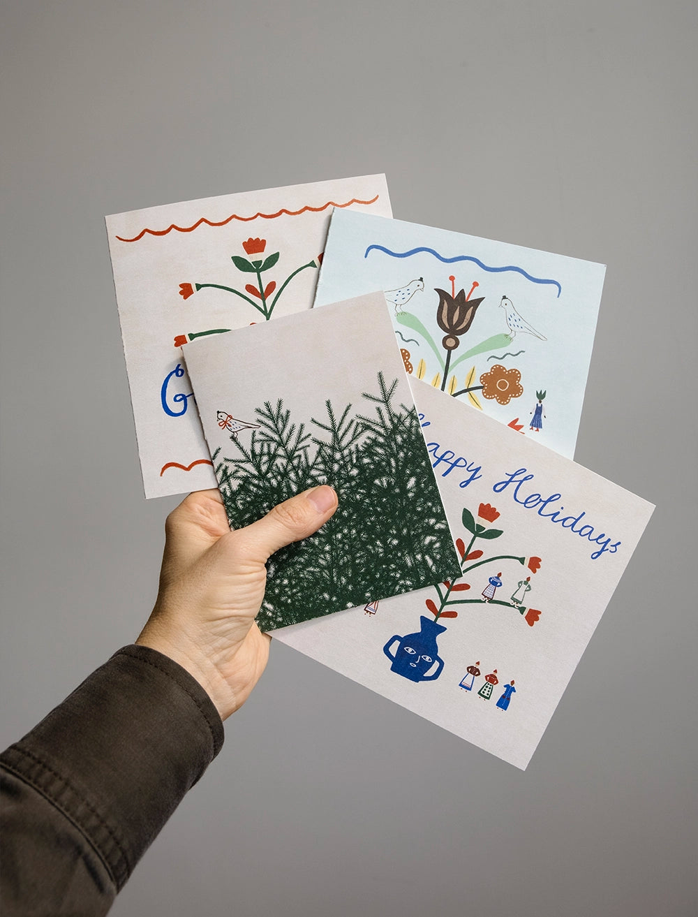 A hand holds four illustrated holiday cards, including the Bow Birdie Card w. Envelope A6 by Fine Little Day, three with festive designs and one featuring green pine branches.