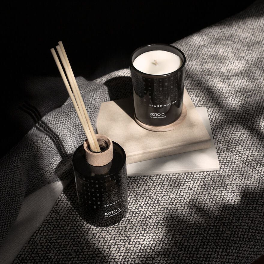 The Skandinavisk Scented Candle Koto 50h, 200g, and a black reed diffuser rest on a beige book atop textured gray fabric in natural light, infusing the space with a subtle Nordic fragrance.