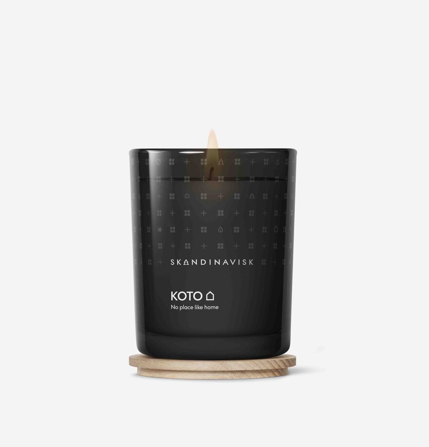 A Skandinavisk Scented Candle Koto 50h, 200g in black glass burns on a round wooden coaster, set against a plain white background.