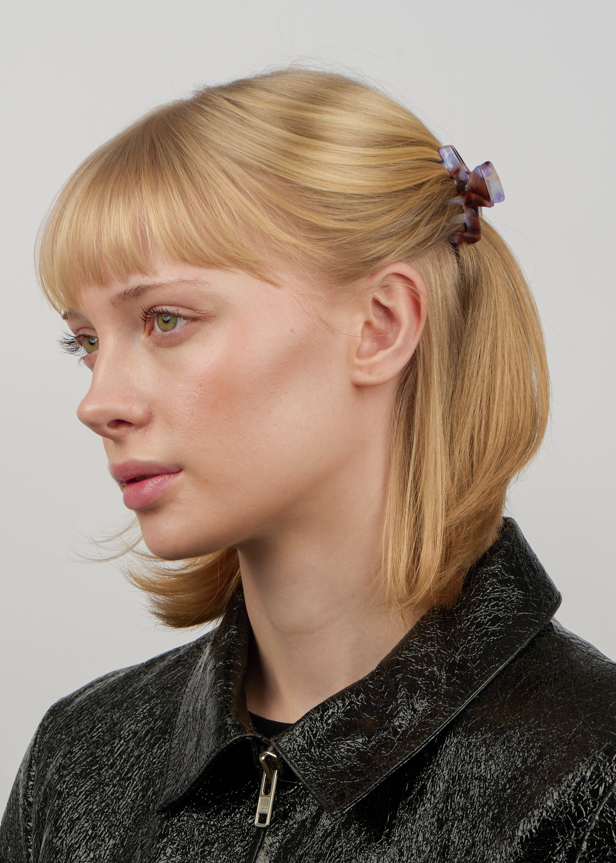 A young woman with blonde hair styled half-up, half-down, secured by the Sui Ava Lea Believe Mini Hair Claw Clip in Sky Blue, wears a shiny black textured jacket with a zipper and looks left against a plain backdrop.