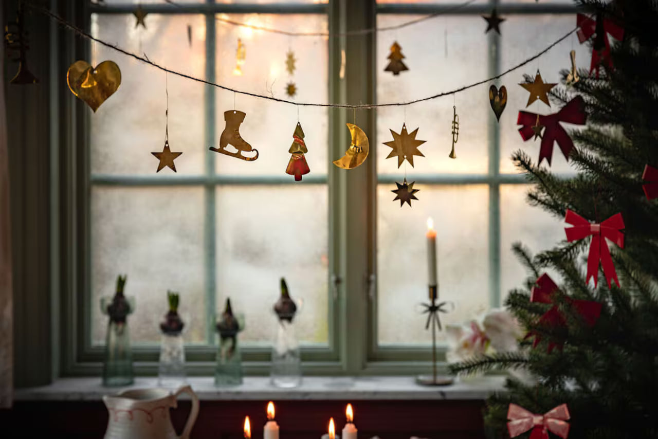The Strömshaga Garland with Ornaments in Brass (2.7m), featuring stars, hearts, a boot, and a tree, hangs by the window; candles light up the table and a decorated tree stands to the right.