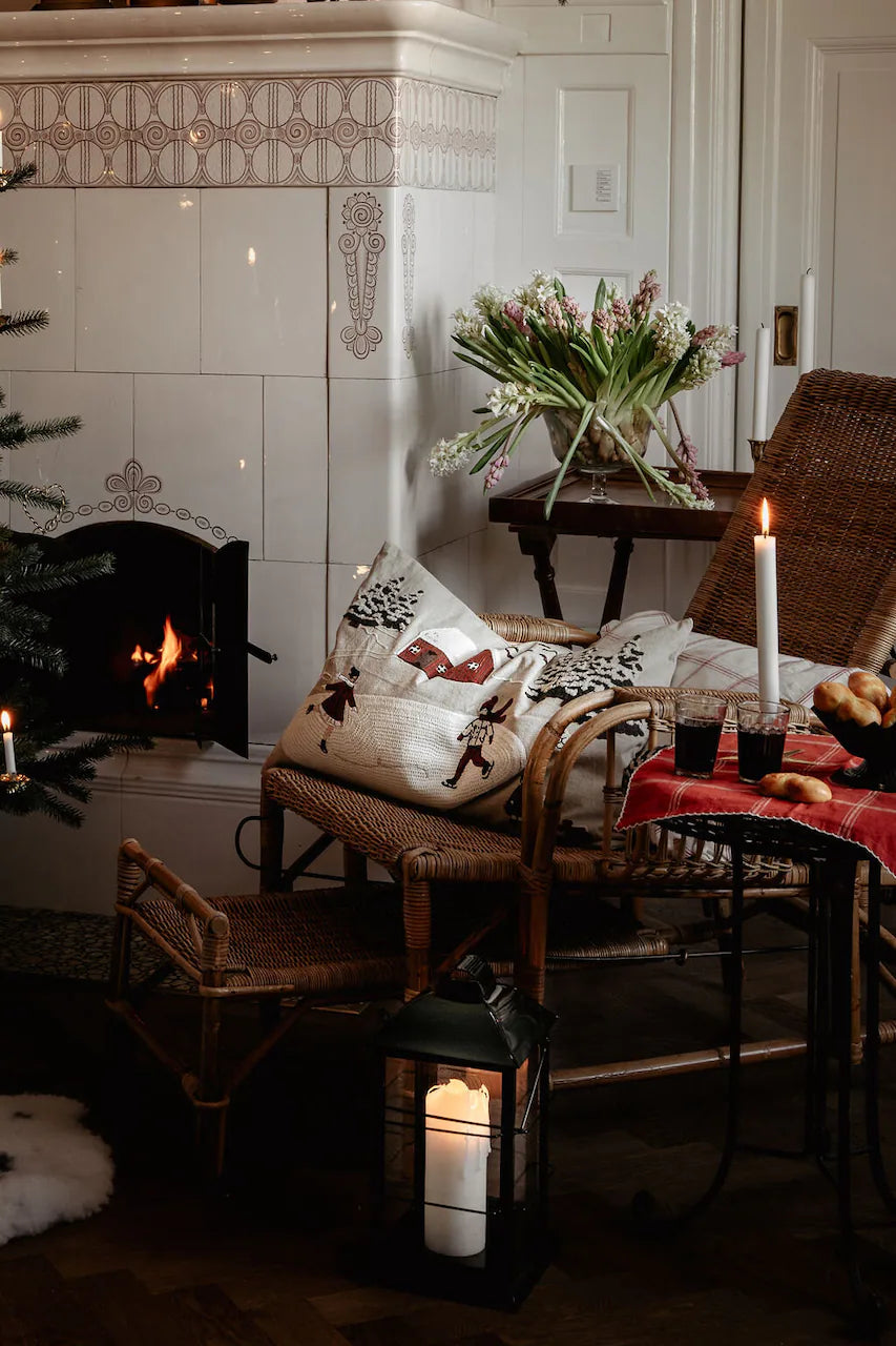 A wicker chair with a Strömshaga Cushion Cover Vinterland Embroidered (40 x 60 cm) sits by a glowing fireplace. On a small table are a candle, flowers, drinks, and pastries, plus a lantern with a candle glowing on the floor.