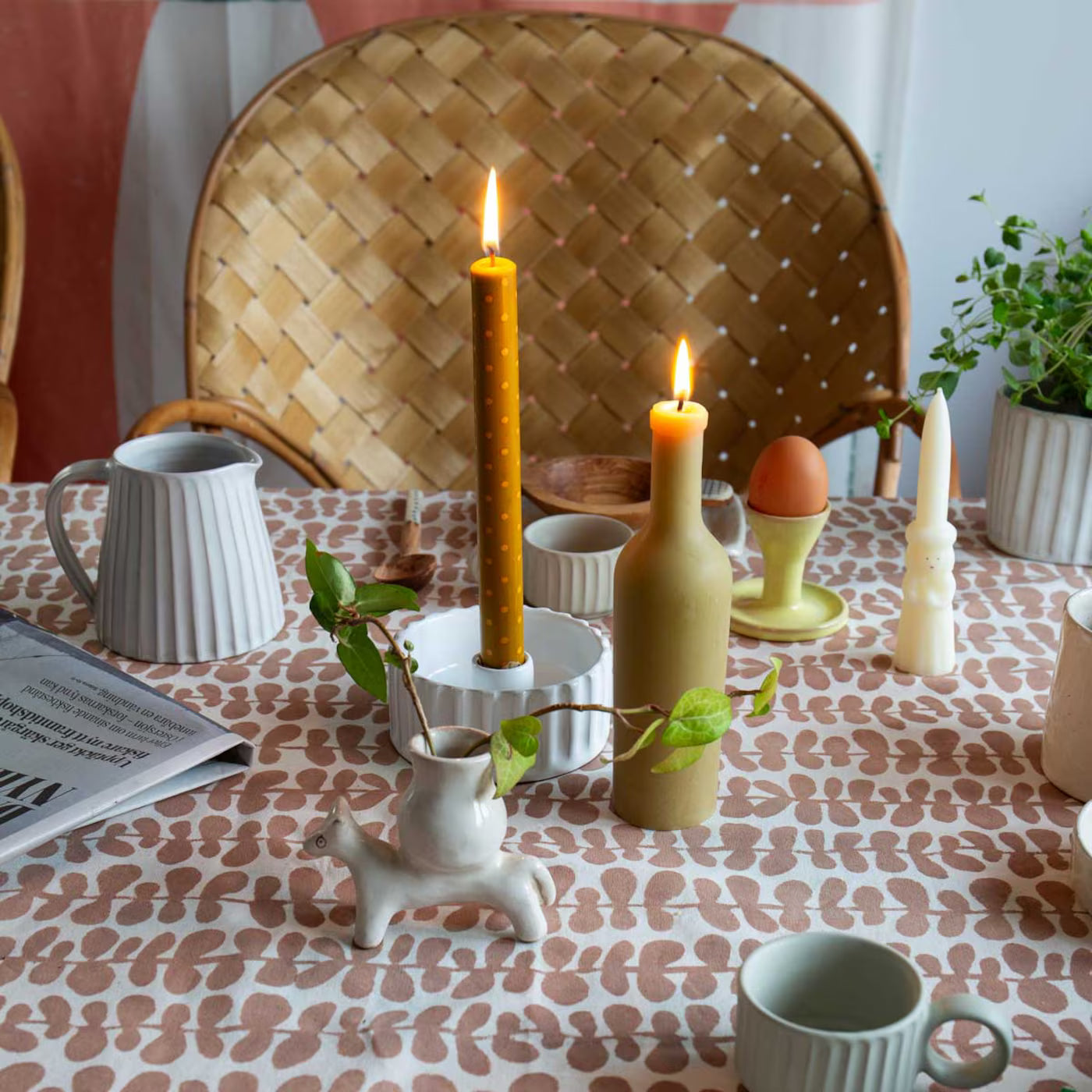 A World of Craft’s handmade Tablecloth Foliage in Pink & Off White (140x260 cm) sets the table, displaying two lit candles, ceramic cups, a plant, a pitcher, an egg holder, animal creamer, newspaper, and woven chairs in the background.