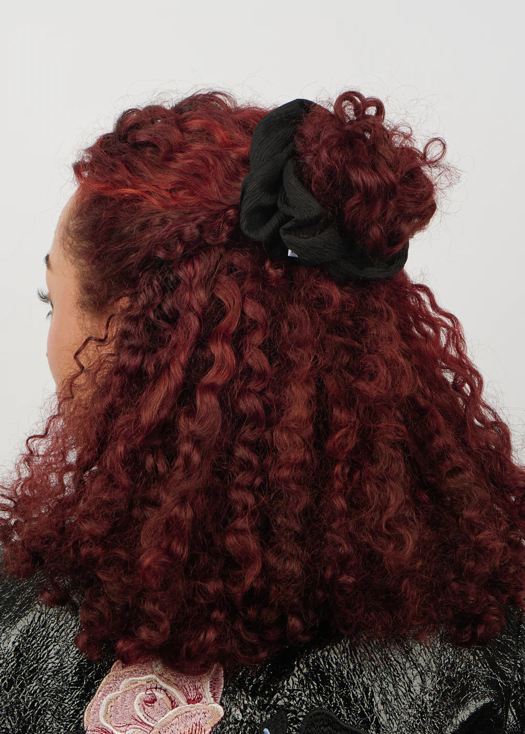A person with curly, burgundy hair styled in a half-up bun secured with the Sui Ava Ea Zig-Zag Scrunchie in Black, facing away from the camera against a plain background.