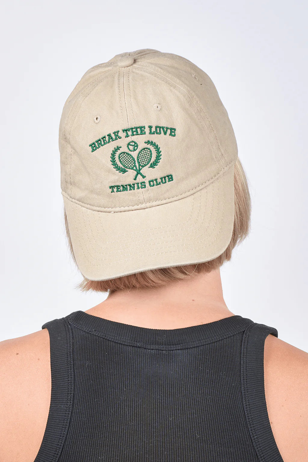 A person wears the Sui Ava Everyday Cap, Tennis Break the Love in Beige—100% cotton, washed beige with green BREAK THE LOVE TENNIS CLUB text above two crossed tennis rackets—shown from behind.