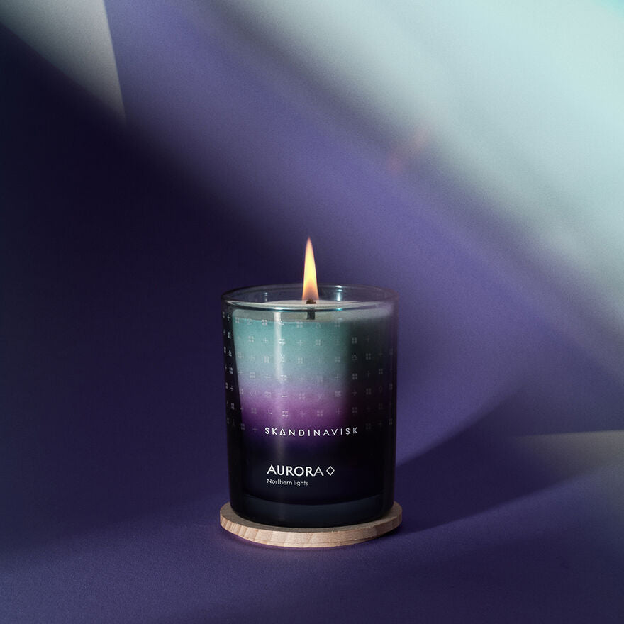 A Skandinavisk Seasonal Aurora Scented Candle 50h, 200g, made from Swedish rapeseed wax, labeled "Northern Lights," sits on a wooden coaster against a gradient blue and purple backdrop.