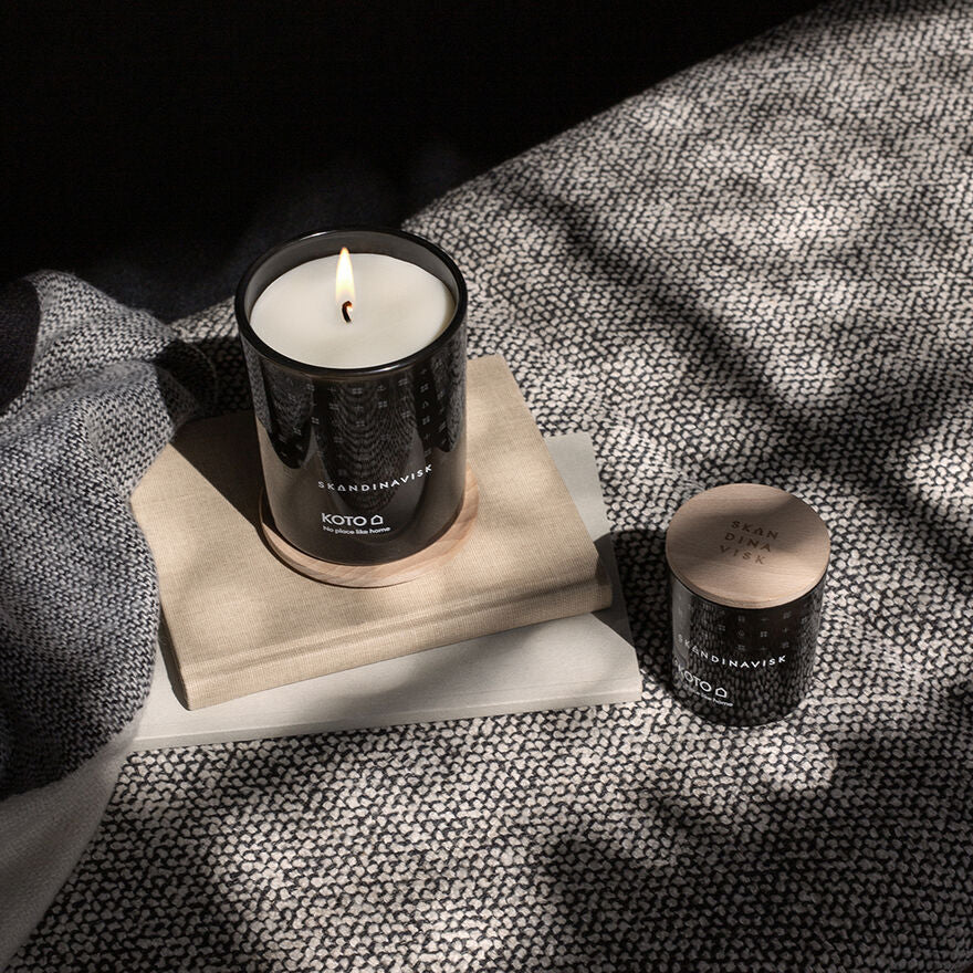 A lit Skandinavisk Mini Scented Candle Koto 20h, 65g in a black glass jar rests on stacked books beside its closed match, both on textured gray fabric, adding Nordic fragrance charm.