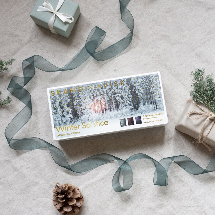 The Skandinavisk Scented Candle Winter Solstice Mini Giftset w. Arktis, Jul & Aurora sits on a beige surface with gifts, green ribbon, pine cone, and greenery—perfect for gifting these Swedish rapeseed wax candles.