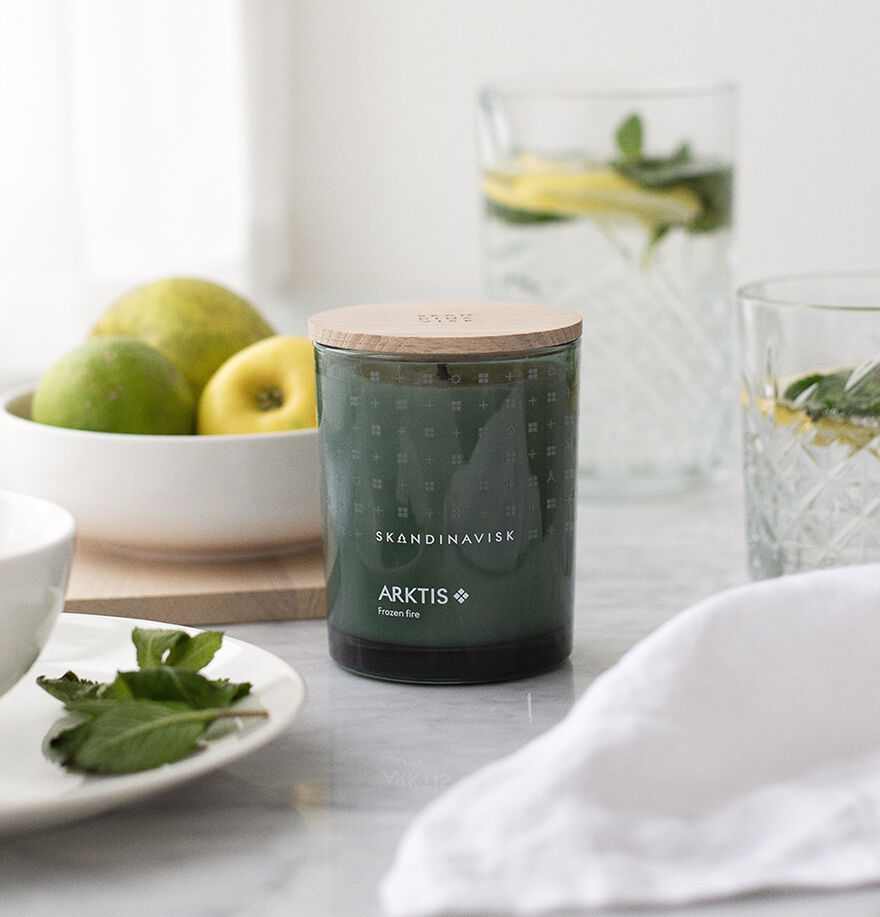 A Skandinavisk Scented Candle Arktis 50h, 200g with a wooden lid rests on marble beside limes and a glass of lemon water, filling the space with an arctic sage fragrance.