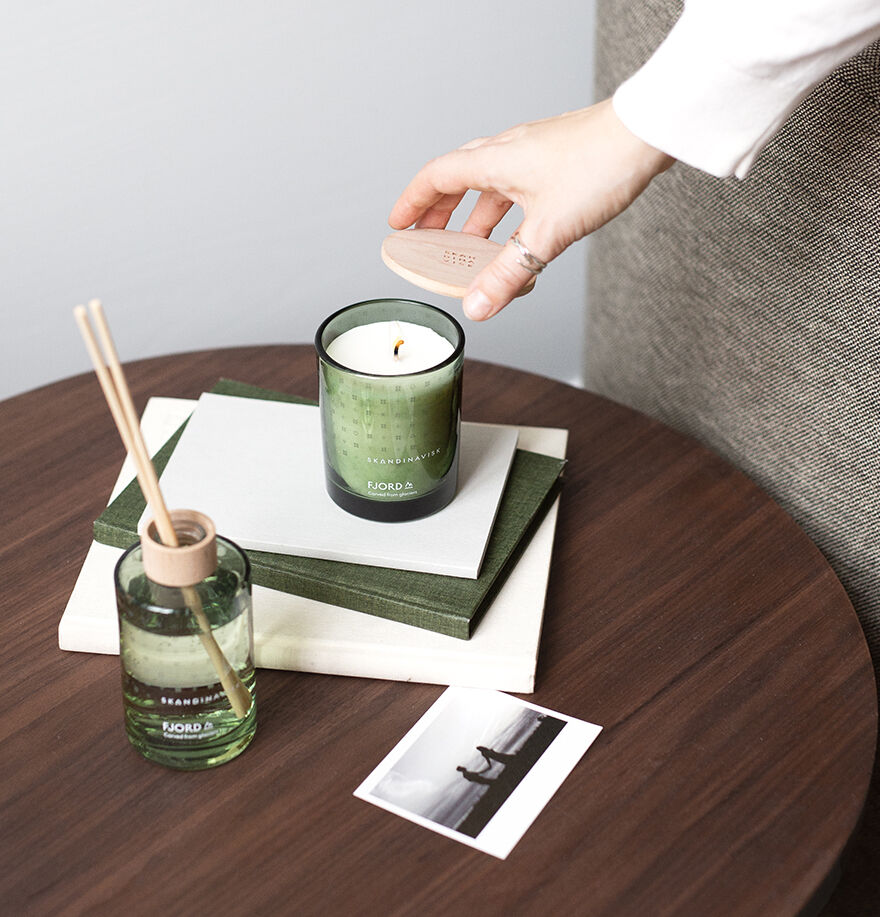 A hand places a wooden lid on the Skandinavisk Scented Candle Fjord 50h, 200g—a green glass jar made with Swedish rapeseed wax—beside a reed diffuser, two books, and a photo on a round wooden table.