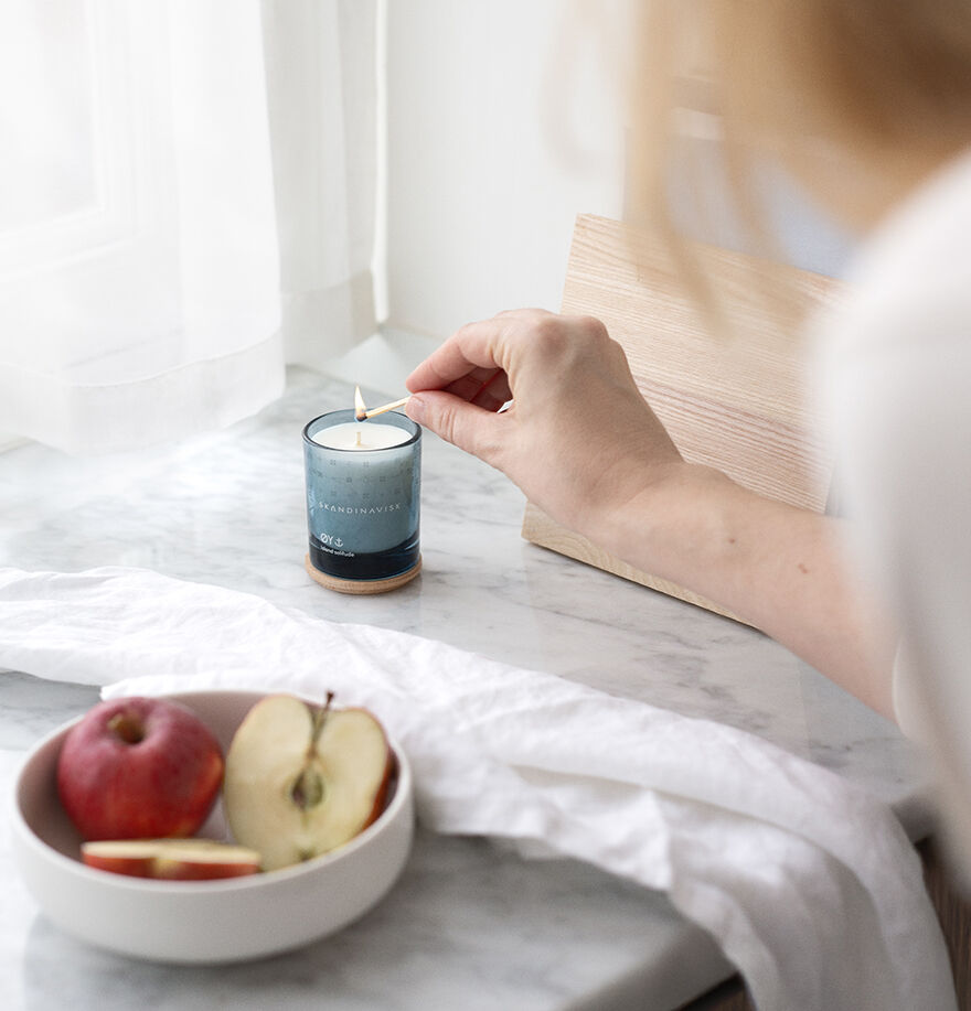 A person lights the Skandinavisk Mini Scented Candle Oy 20h, 65g made from Swedish rapeseed wax on a marble countertop beside a bowl with a whole and sliced apple.