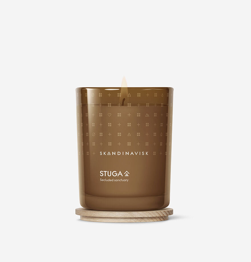A lit Skandinavisk Seasonal Stuga Scented Candle 50h, 200g in brown glass sits on a round wooden coaster, evoking woodland mosses against a plain background.