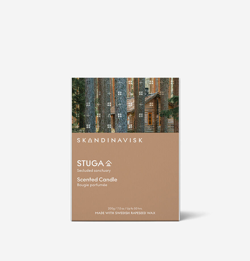 The Skandinavisk Seasonal Stuga Scented Candle 50h, 200g features a wooden cabin among trees, labeled "Secluded sanctuary," and is infused with notes of birch bark and woodland mosses.