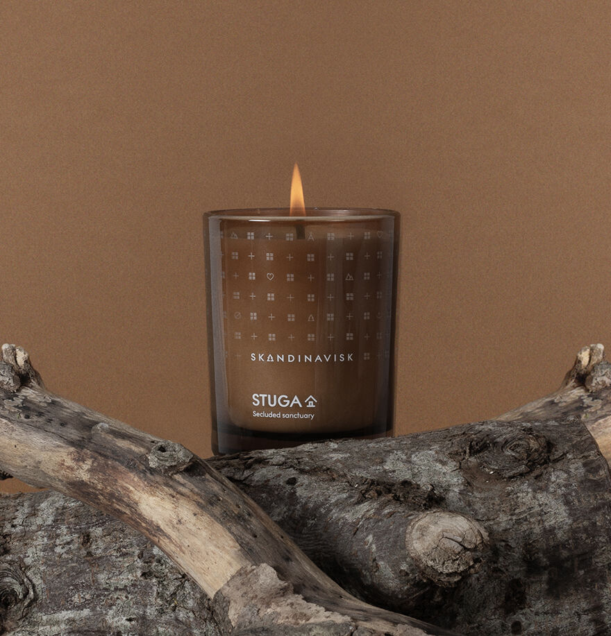 A lit Skandinavisk Seasonal Stuga Scented Candle (200g, 50h) sits on driftwood against a brown background, filling the air with the aroma of woodland mosses.